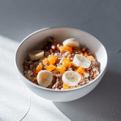 Healthy raw muesli with delicious oatmeal, dried apricots, raisins, walnuts and banana in a white bowl on a white table with sun rays in the morning