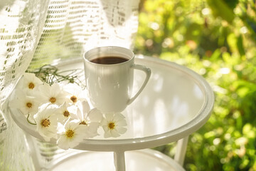 Beautiful porcelain coffee cup with white flowers on glass table in summer garden in sunlight. Top view. Copy space. Summer drinks concept.