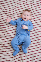 Happy baby boy in blue clothes is lying on a striped bedspread, top view. Smiling child toddler at age six months in red bed, above