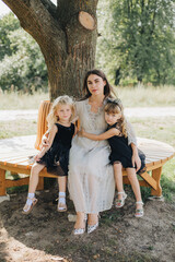 mother and two daughters sitting on a bench under a tree. Woman in a beautiful beige dress. Little girl in a black dress. Mother's Day. Mother's love for her child. Family walks in the park. Twin girl