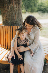 mom and daughter sitting on a bench under a tree. Woman in a beautiful beige dress. Little girl in a black dress. Mother's Day. Mother's love for her child. Family walks in the park