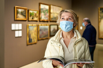 elderly woman in mask protecting against covid examines paintings on display in hall of art museum