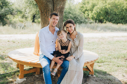 Man And Woman With A Child Sitting On A Bench Under A Tree. Happy Young Couple With Daughter In A Black Dress. Spend Time With Family In The Park