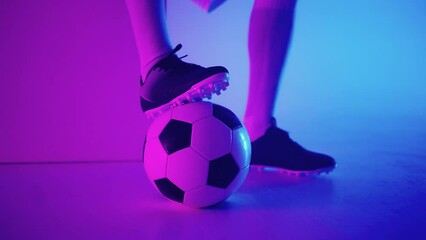 Close-up of the foot of a professional black football player standing on the ball in slow motion in the blue-red neon light of the studio. Brazilian football player foot on the ball to pose