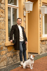 A man walking in the city square with a dog. Jack Russell terrier on a leash near the building.