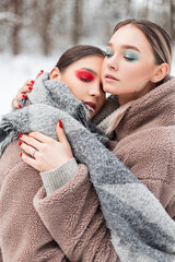 Two stylish beautiful young girls with creative color makeup in fashionable winter clothes with a fur coat and a scarf are hugging outdoors in the forest with snow