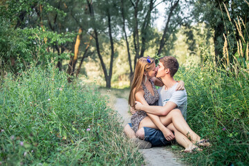 Young happy couple kissing while sitting on a path among the grasses in the summer at sunset.