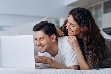 Joyful young couple of wife and brunet husband discusses chosen online purchases at sell-out via laptop lying on soft sofa in living room