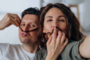 Happy newlyweds play and pose making mustaches with curly brunette hair and kiss smiling broadly for selfies via smartphone closeup