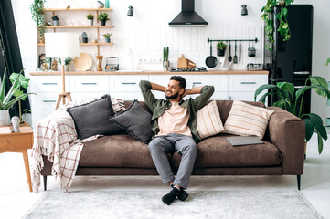 Satisfied relaxed young indian man chilling on sofa in living room, smiling. Handsome arabian guy...