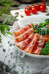 Sliced layers of beef, parma ham with cherry tomatoes on arugula leaves, seasoned with balsamic sauce in a plate on a gray stone table. Restaurant Menu
