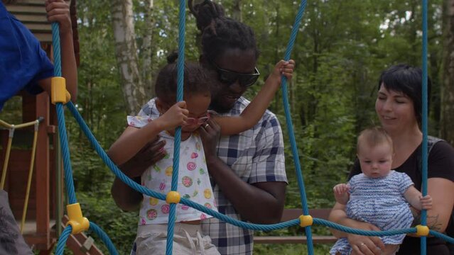 A father helps his daughter overcome an obstacle made of ropes in a playground. Communication between adults on the topic of raising children.