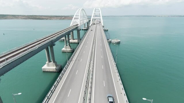 Aerial view along the long beautiful bridge above turquoise sea. Action. Flying above bending bridge with driving cars.