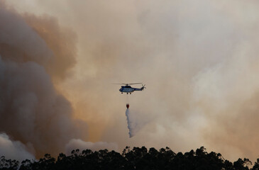 A helicopter flies by discharging water amid clouds of smoke from a forest fire