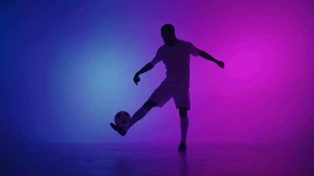 Black man a player juggles football ball in a dark studio with neon lights on the floor and red and blue lighting effects in slow motion. African professional football soccer player