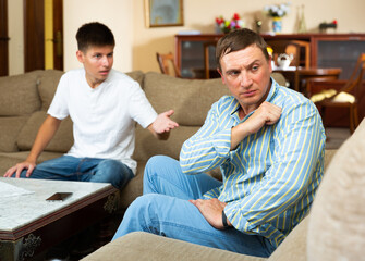 Upset father sitting while quarreling with his teenage son in domestic interior