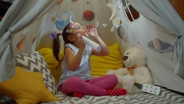 Portrait of a cheerful cute preschool girl watching the planets through a paper telescope, sitting at home in a tent. Smart little girl is playing the explorer of space and planets.