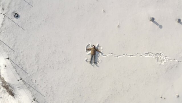 Handsome young man making snow angels and playing in snow in an empty city park during winter season. Overhead aerial top view from the drone. Snow angel on white covered winter snowy field.