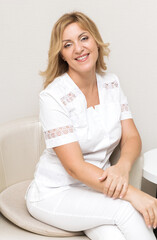 A beautiful beautician doctor in a white uniform smiles, looks into the camera and poses while sitting in a chair in her office.