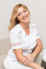 Naklejka premium A beautiful beautician doctor in a white uniform smiles, looks into the camera and poses while sitting in a chair in her office.