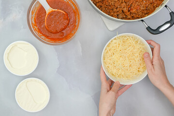 Parmesan cheese close up in a bowl, and some ingredients for beef lasagna recipe close up on kitchen table, flat lay, woman hands