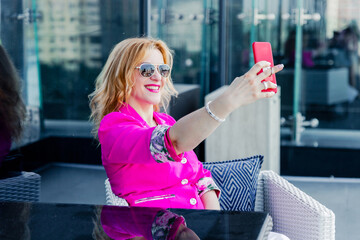A successful woman entrepreneur in a magenta jacket makes a selfie on the terrace of a cafe in a business center.