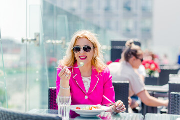 Beautiful woman in a bright magenta jacket sits on a summer terrace and eats a fresh vegetable salad