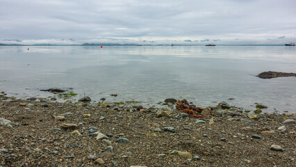 A ship is sailing on the smooth, calm surface of the Pacific Ocean. On the horizon is a mountain range hidden in the clouds. In the foreground is a pebble beach. Petropavlovsk-Kamchatsky