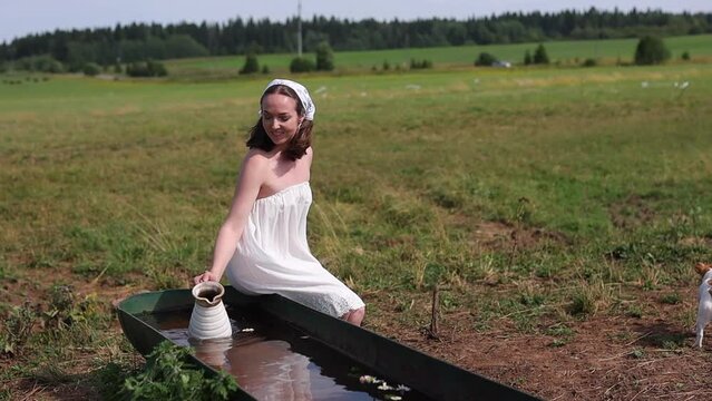 A girl in a headscarf and a light summer dress with a jug. A girl draws water into a jug and pours it out. lively wildflowers floating on the water. daisies and yellow flowers in a bathtub on a summer