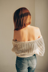 sexy young woman with bare shoulders in knitted cardigan, jeans stands freely with her back in sunlight bright room. free natural girl. vertically, selective focus