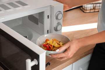 Young woman putting bowl with delicious food into modern microwave oven on wooden counter