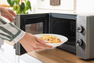 Woman putting plate of rice with vegetables into microwave oven in kitchen