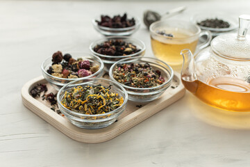 Different varieties of herbal and fruit tea in glass bowls next to the kettle filled with a hot drink and a cup. Close-up and selective focus. Vegan drinks. Medicinal herbal collection. 