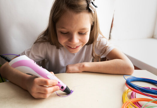 A 7-year-old girl with long hair and a white T-shirt is sitting at a table making figures with a pink 3-d pen. Toys made of plastic wire.