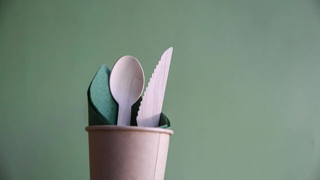 Group of wooden disposable cutlery (forks, spoons and knifes) falls in brown paper cup with green napkin standing against green background. 4K resolution video. Environmental conservation theme.