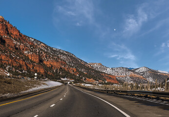 View of highway through red rock formations in Colorado in winter from moving car; moon rising