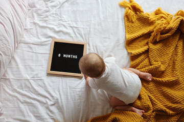 Caucasian blonde baby eight months old lying on cozy bed with yellow knitted blanket at home. Kid wearing white clothing 