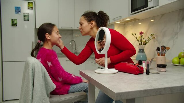 Cheerful mother applying mascara to preteen girl at home. Reflection of smiling femele child in vanity mirror. Beautiful parent spending time together with adorable daughter indoors. Kid's cosmetics