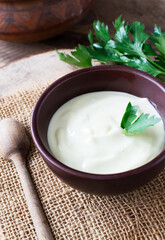 Homemade mayonnaise in a brown bowl with a sprig of green parsley on a wooden table. The concept of proper nutrition. Rustic. Vertical orientation.