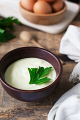 Homemade mayonnaise in a brown bowl with a sprig of green parsley and eggs on a wooden table. The concept of proper nutrition. Rustic. Vertical orientation.