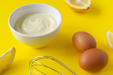 Homemade mayonnaise in a white bowl with eggs and lemon slices and a whisk on a yellow background. The concept of proper nutrition. Creative. Horizontal orientation.