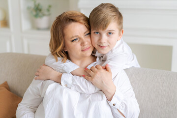 Happy young mom hugs her toddler son, relaxing on sofa at home. Cute toddler boy and smiling mother approach each other, hug. Adorable little baby boy and mom enjoying moments of love and affection.