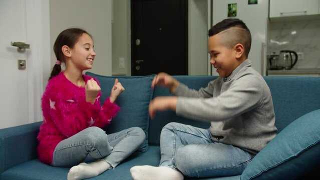 Charming preteen girl in pink pullover teaching cute black boy with yes gesture while sitting on couch indoors. Multiethnic boy and girl giving high five, smiling, laughing together at home