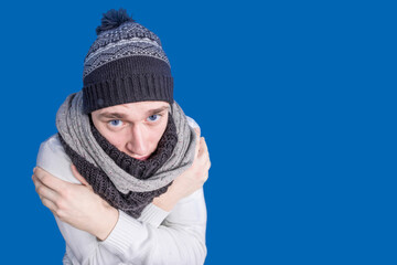 A man in a fur hat and scarf crossed his arms from the cold on a blue background. Freezing person. Warmly. Winter. Coldly