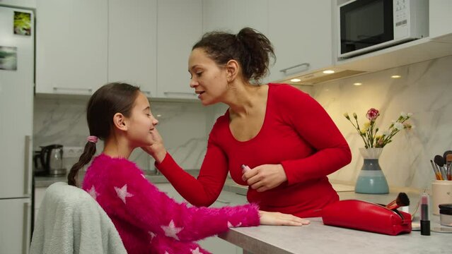 Caring mother teaching lovely daughter putting on makeup on face skin indoors. Adult black woman applying mascara to preteen girl eyelashes, using cosmetics at home. Family spending day together