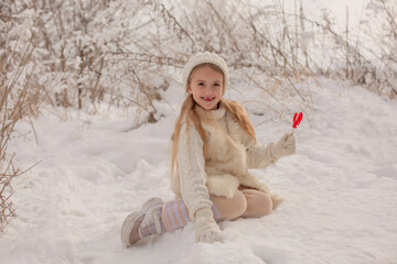 a blonde girl in a white knitted sweater and hat is sitting in the snow in a snowy winter park with a lollipop in the form of a cockerel in her hands