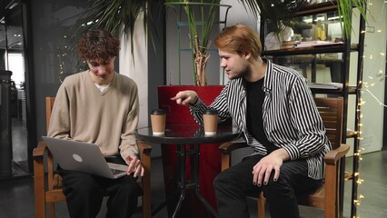 Two gay guys on a date are sitting in a coffee shop, drinking coffee and looking at something on a laptop. LGBT