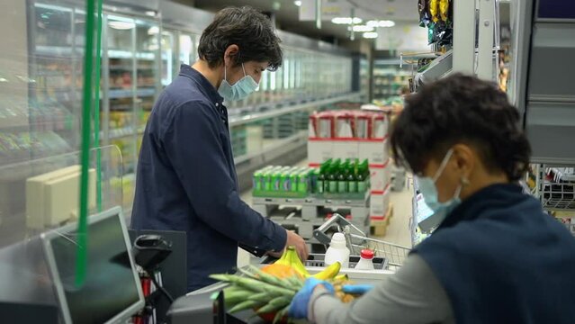 Male Consumer Making Purchases At Supermarket Checkout And Cashier Selling Spbd. Closeup View Of Young Man Puts Fresh Fruit On Tape And Stands At Counter, Female Worker Scans And Makes Sale At