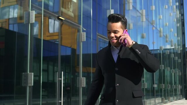 Portrait of young businessman talking on phone and walking in modern city spbd. Closeup view of American man looks with smile and has mobile call, discusses business and walks in town on autumn day