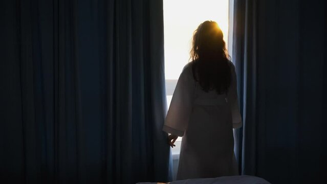 Silhouette woman in white bathrobe opens curtains in dark badroom after waking up looking outside large panoramic window enjoying sunrise in early morning